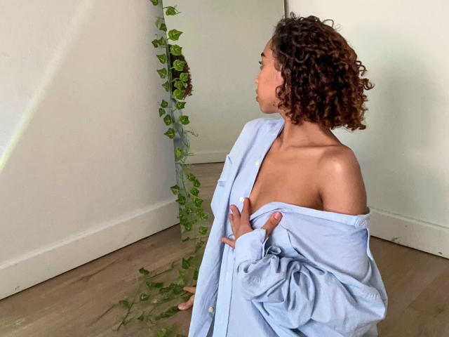 AngelitaFlor, con i capelli castano ricci, la pelle color nocciola e un sedere piccolo, indossa una camicia blu portata fuori dalla spalla, toccandosi il seno, in piedi su un pavimento di legno con delle viti verdi..
