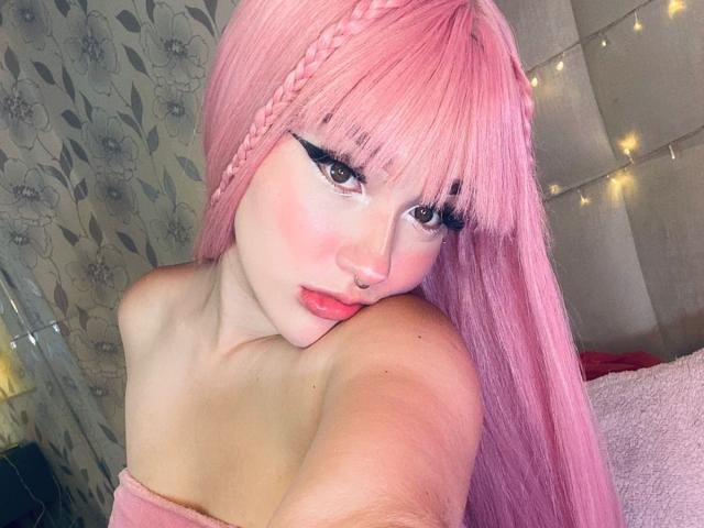 CristySanz with pink hair, braided front, black eyeliner, pink blush, nose ring, and red lipstick, looking at camera, topless, showing shoulders, with a pouty expression.