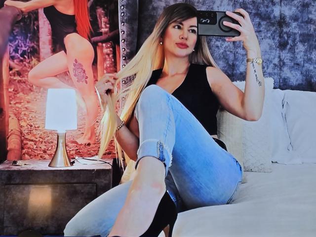 Leared with long blonde hair, black top, and ripped blue jeans, taking a mirror selfie, showing her tits and ass, sitting on a bed with a lamp and poster.