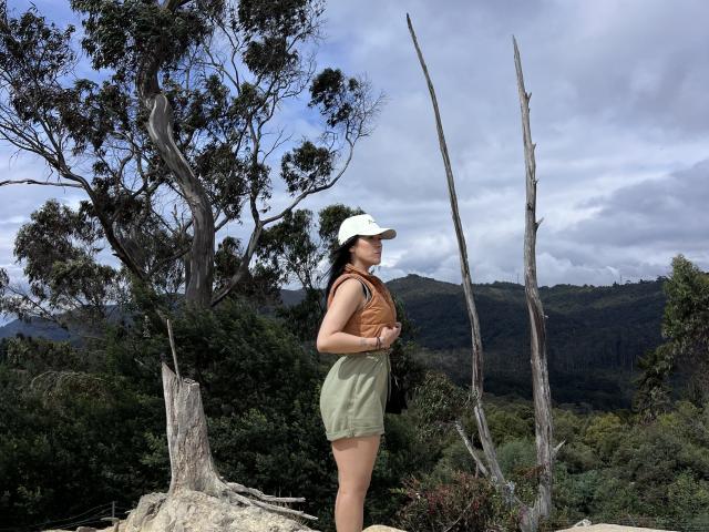 NicoColeman stands in a forest, wearing a white cap, brown vest, and green shorts, with a backpack, showing her ass and looking away from the camera.