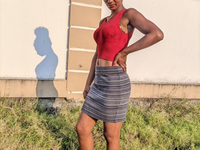 Sanda with dark skin, red tank top showing her tits, and a striped skirt, standing on grass, shadow on wall, hands on hips, midday sunlight.