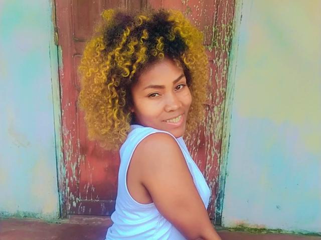 Avax69 with curly yellow and brown hair, smiling at the camera, wearing a white sleeveless top, showing her back and butt, standing in front of a weathered red door.