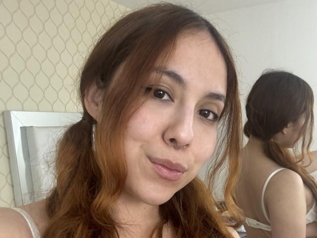 AmberlStha, com maria-chiquinhas castanhas, um pequeno piercing no nariz e um sorriso discreto, olhando para si mesma em um espelho, que reflete sua imagem, mostrando sua camiseta branca e seus olhos castanhos..
