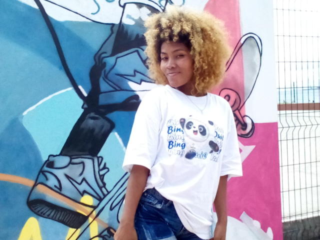 Avax69 with curly blonde hair, wearing a white t-shirt and blue jeans, standing in front of colorful graffiti, smiling slightly. Avax69's t-shirt has panda illustrations and text.