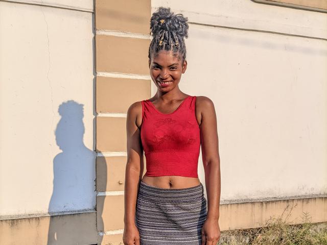 Sanda has dark skin, small tits in a red crop top, and a striped skirt, standing with a smile, gray braided bun, shadow on wall, sunlight.