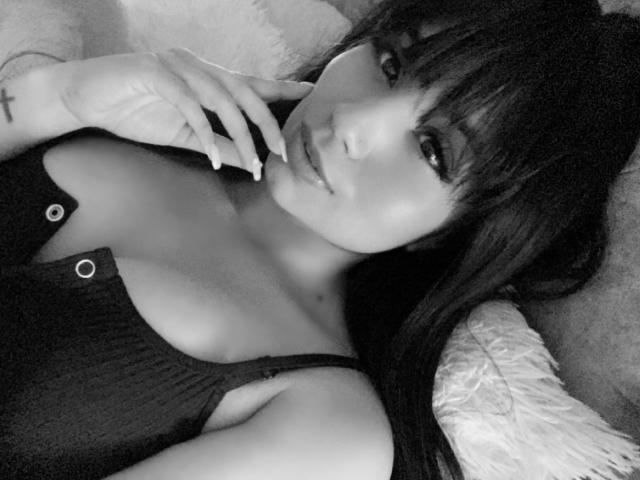 Black-and-white selfie of KattyWattson with dark hair, bangs, and seductive eyes, lying on a fluffy pillow, wearing a ribbed, buttoned-up top, showing her tits.