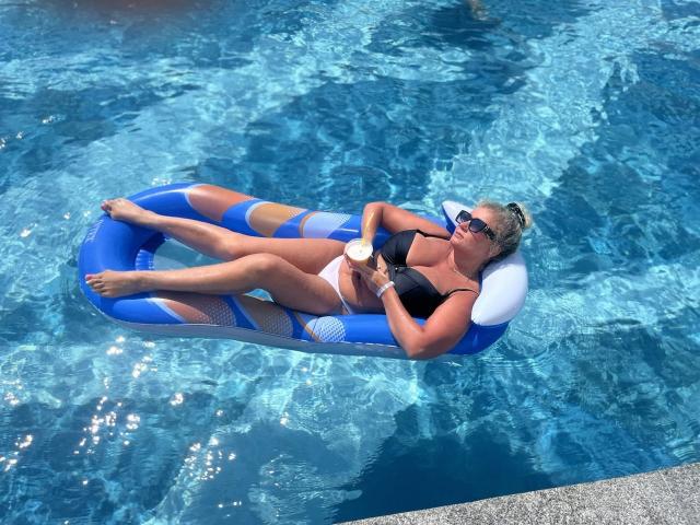 Marmeladka69, con la pelle chiara, i capelli biondi raccolti in uno chignon, un bikini nero, occhiali da sole, sdraiata su un materassino gonfiabile blu, tiene in mano una bevanda bianca in una piscina. Il suo sedere è sopra l'acqua..