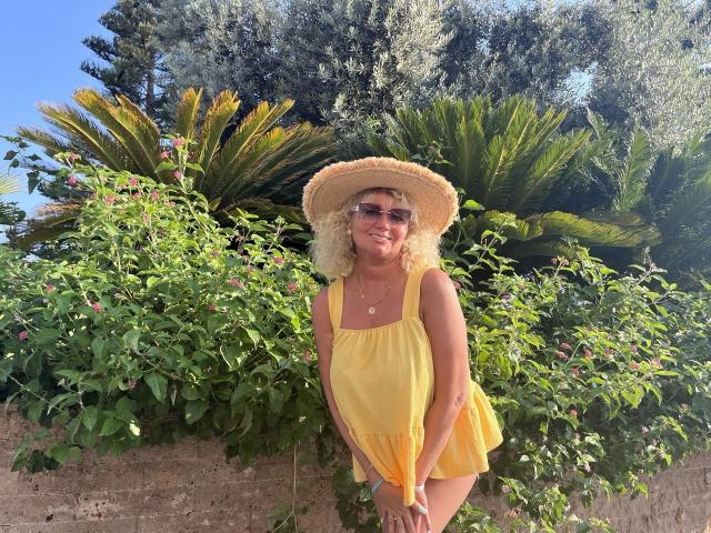 Marmeladka69 con un vestito giallo e un cappello di paglia, piegata in avanti con un sorriso, che indossa occhiali da sole, con capelli biondi ricci, che mostra i suoi seni sotto il vestito. Giardino illuminato dal sole..