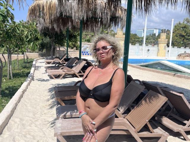 Marmeladka69, con i capelli biondi ricci, indossa un reggiseno nero e mutandine abbinate, ed è in piedi di fronte a lettini di legno, con le mani giunte, occhiali da sole, in un'area esterna soleggiata con una piscina, con un'espressione seria..