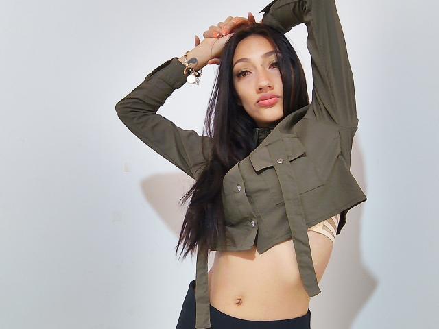 KiriamGomez, con lunghi capelli neri, indossa una giacca verde in stile militare e ha le braccia alzate, mostrando il suo seno piccolo e il ventre piatto. Ha addosso un reggiseno beige e pantaloni neri..