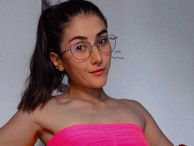 AyanaTessy with pink strapless top, glasses, and ponytail, showing her tits and a subtle smile, in a selfie. She has light skin and a cute, confident look.