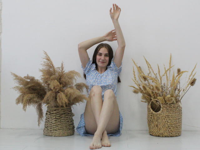 NikaStark with arms raised, wearing a blue floral dress, sitting on white floor between two wicker baskets with dried pampas grass, smiling, barefoot, legs crossed.