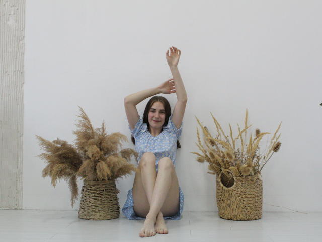 NikaStark sits on floor, arms raised, wearing blue floral dress, legs crossed, barefoot, flanked by two wicker baskets of dried pampas grass, smiling.