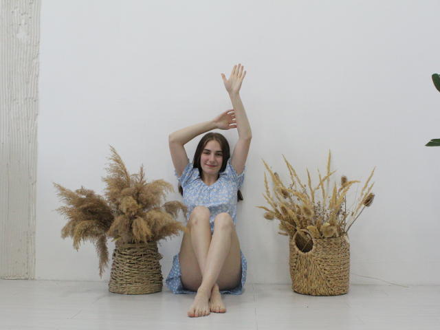 NikaStark sits barefoot with arms raised, wearing a blue floral dress, between two wicker baskets of pampas grass. She has a cute smile and small tits.