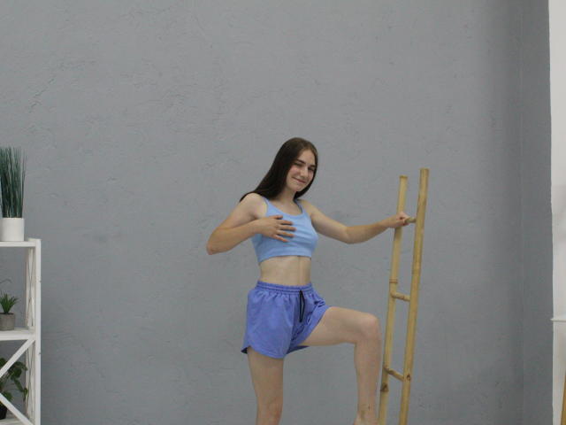 NikaStark with medium tits, blue sports bra, and purple shorts, standing on a wooden ladder, holding her left breast with right hand, smiling, long brown hair, gray wall.