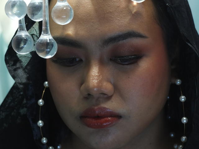 SweetBigCockForYou with dark skin, red lipstick, and pearl earrings, looking down with a serious expression. Glass beads hang over her head, black lace covering it.