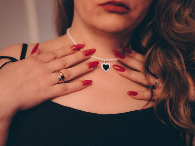 AdelaideRosabel's hands with red nails, black dress, pearl necklace, and heart pendant, wearing gold rings, touching chest, wavy brown hair, red lipstick.