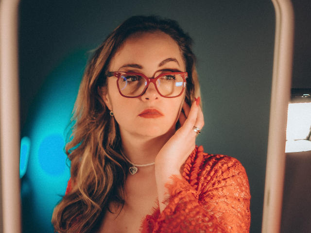AdelaideRosabel con gafas rojas, blusa de encaje naranja, cabello rubio ondulado, mano tocando la mejilla, collar con forma de corazón, expresión seria, reflejo en un espejo, iluminación suave, fondo azul y blanco sutil..