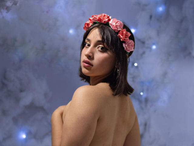 VickyButerfly, topless with pink rose headband, medium tits, short dark hair, looking over shoulder, soft light, blue-gray background, subtle bokeh, bare back, gentle expression, small gold earring.