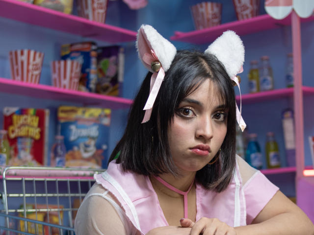VickyButerfly with white furry cat ears and pink ribbons, wearing a pink and white sheer top, pouty lips, and short black hair, looking serious in a brightly lit store.