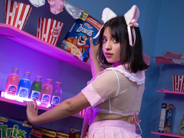 VickyButerfly with cat ears, pink bow, sheer pink top, pink skirt, reaching for neon blue soda bottles, looking back with a cute, playful expression.