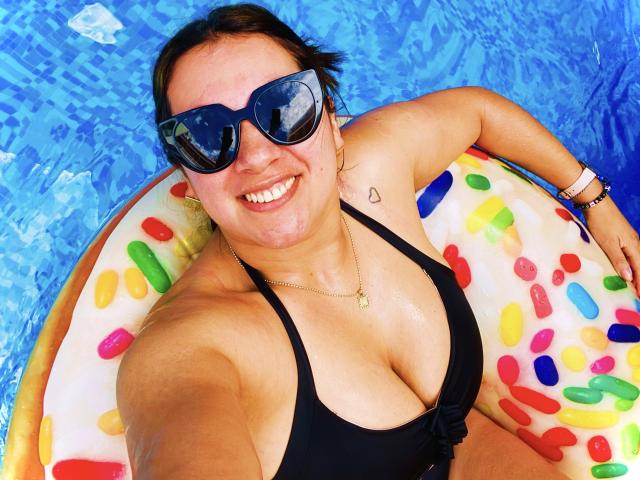 AnnaPaulie with big tits, wearing black bikini top, sunglasses, and bracelet, lying in blue pool on colorful inflatable ring, smiling at camera. Small tattoo on right shoulder.