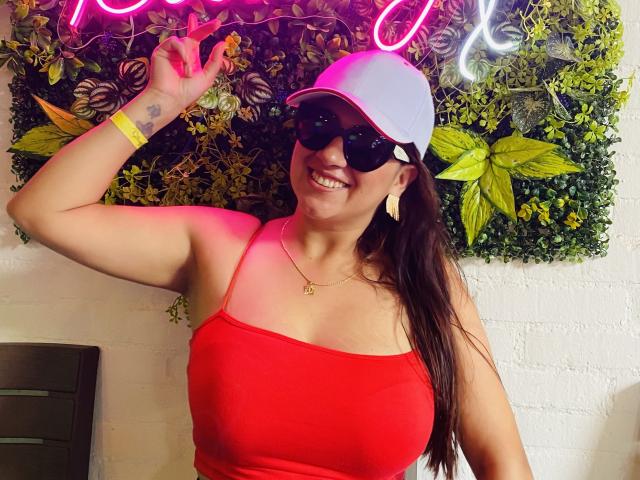 AnnaPaulie with big tits, wearing a red tank top, white cap, black sunglasses, smiling, holding up a peace sign, green wristband, yellow necklace, and yellow earrings.