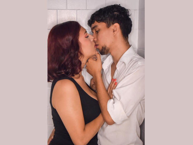 SpecialFriendsBenefits with red hair and black tank top kissing SpecialFriendsBenefits with curly hair and white shirt, holding face and chest. Her hand with red nails on his neck.