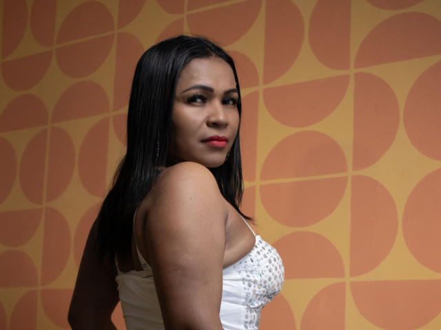LouiseNails with dark hair, red lipstick, white dress with sparkly straps, looking over shoulder, showing her tits, standing against patterned wall.