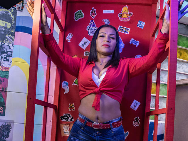 LouiseNails, with medium tits, wearing a tied red shirt, blue jeans, and a belt, standing with arms up, in a red, sticker-covered frame, looking confident.