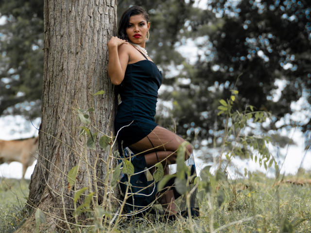 MarceBetancur leaning against a tree, wearing a black dress, brown stockings, and black shoes, with her hand on the trunk, looking at the camera with red lipstick.