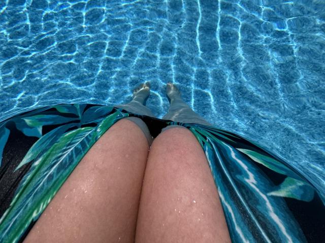 Barbarica's thighs in a green and blue floral swimsuit, submerged in a clear blue pool, with water ripples and light reflections on her pale skin.
