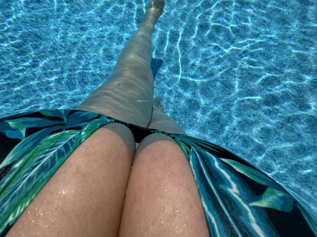 Barbarica's thighs in a green and blue leaf-patterned swim dress, submerged in clear blue water, with her legs extending upwards. Her thighs are slightly wet and visible.