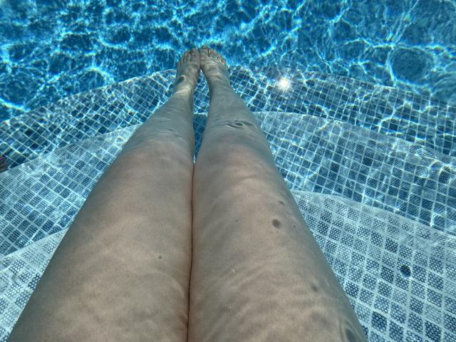 Las piernas de Barbarica en una piscina, mostrando su piel clara y ondulada con pequeñas pecas, sumergidas en agua azul cristalina, con los pies tocándose, las piernas ligeramente separadas, y una cubierta de malla blanca para la piscina..