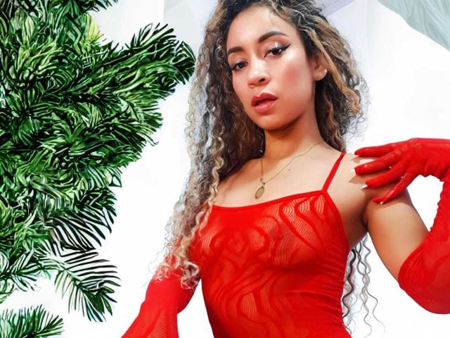 KylieJen with curly hair in a red, sheer, lace-up top and matching red gloves, showing her small, perky tits, and a gold necklace, against green palm leaves.