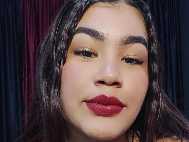 O rosto de AmyXPlata, com cabelos castanhos ondulados, sobrancelhas escuras e batom vermelho, sorri suavemente. Seus olhos têm cílios longos e grossos, e sua pele é clara e lisa..