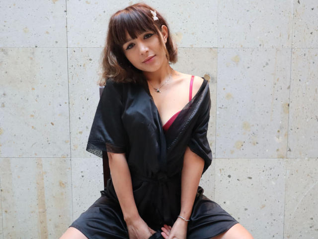 Katy2Kiss-hot with brown hair, pink bra, and black silk robe, sitting with hands on knees, looking at camera, wearing a hair clip and bracelet.