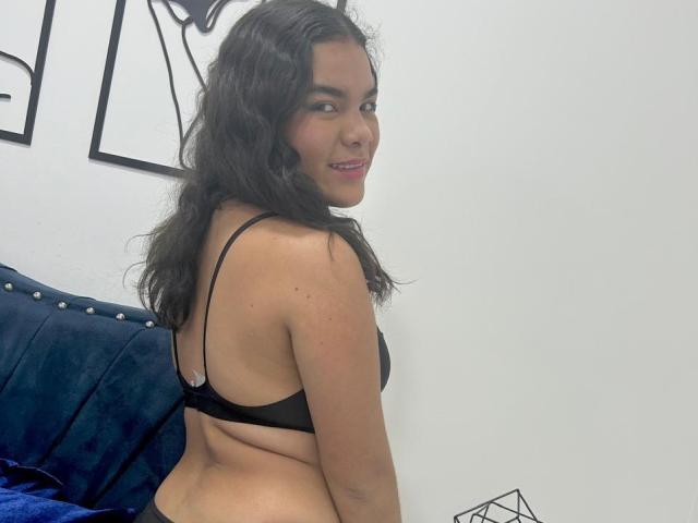 EstrellaBoots with wavy black hair, smiling, wearing a black bra, showing her back and butt, sitting on a blue studded couch, looking over her shoulder.