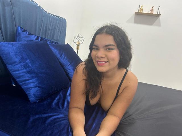 EstrellaBoots, with medium brown skin and long curly black hair, is lying on a bed with blue velvet pillows, showing her small tits in a black spaghetti strap top.