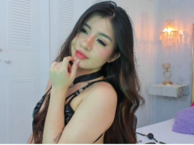 SaraLens with long brown hair, wearing black lingerie, has her finger near her red lips, smiling. Her tits are visible, and she's touching her chin. Bedroom setting.