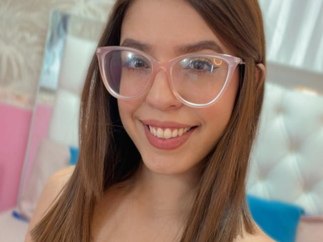 NatyBakers, con cabello castaño, usando gafas transparentes rosas, sonriendo mostrando los dientes, con labios rosados y piel clara. Sus senos no son visibles. Está en un ambiente interior con luz suave..