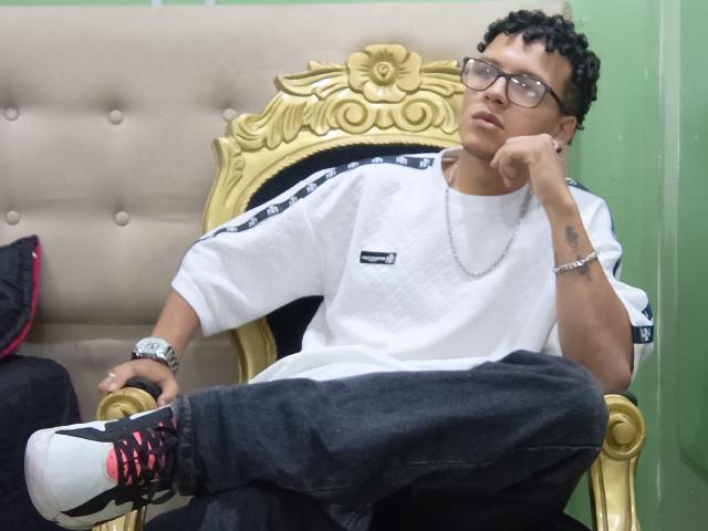 BonnyBonss with curly black hair, glasses, white shirt, black jeans, and red sneakers, sitting on a gold throne, hand on chin, looking bored, silver chain, green wall.