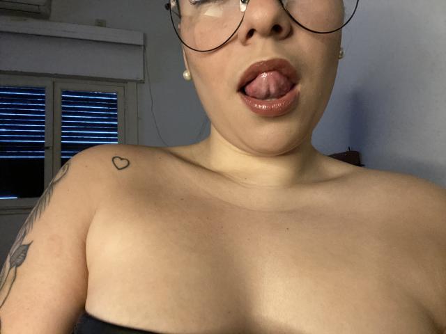 AlexaLip has light skin, glasses, and is sticking out her glossy pink tongue. She's topless with small tits, a heart tattoo on her shoulder, and a tattoo on her arm.