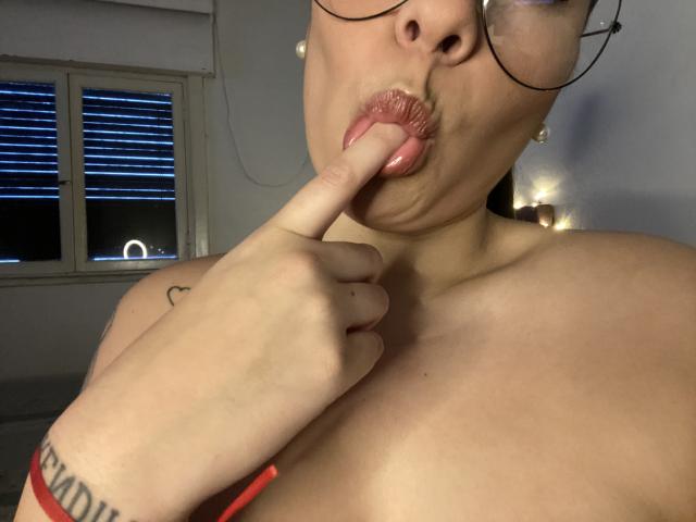 AlexaLip with her finger in her pink, glossy lips, wearing glasses, topless, showing her tits and a tattoo on her wrist. Night outside the window.