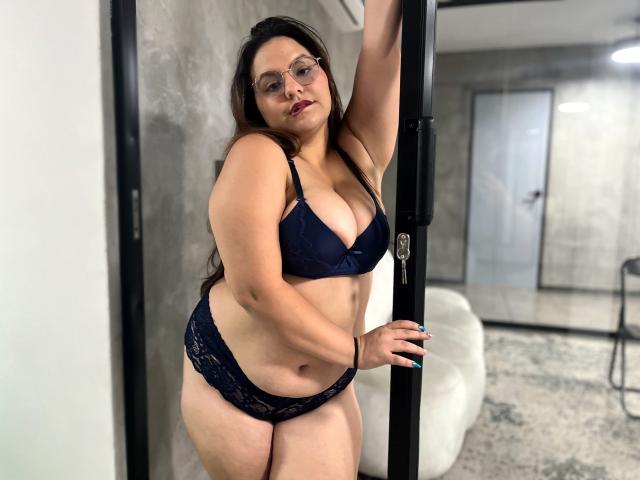 DanielleLopez with big boobs in dark lace bra and panties, standing in a doorway, arm raised, wearing glasses, looking at camera, slightly curvy, light skin, dark hair.