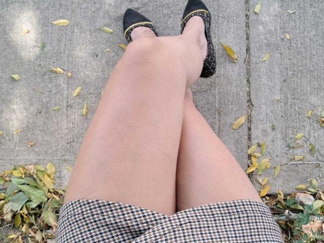 LaurellCharming's legs crossed at the knees, wearing a black and white houndstooth skirt and black shoes with gold accents, on a concrete surface with scattered leaves.