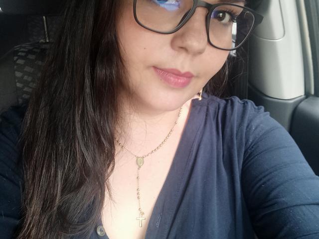 MilfChel with long black hair, wearing glasses and a navy blue shirt, has pink lips and a gold necklace, sitting in a car. Her tits are small and her pussy is unseen.