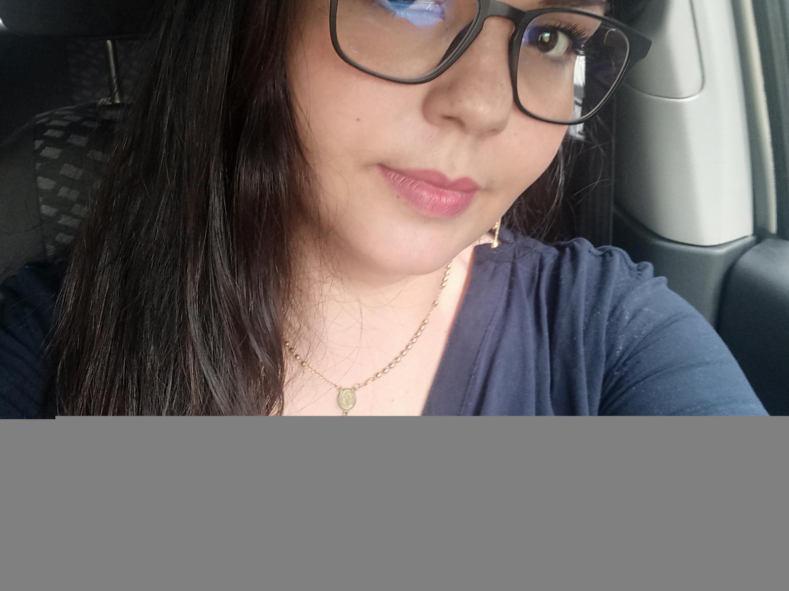 MilfChel with long black hair, wearing glasses and a navy blue shirt, has pink lips and a gold necklace, sitting in a car. Her tits are small and her pussy is unseen.