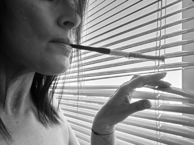 ClementineFoxy with a cigarette in her mouth, her fingers partially holding blinds, in a black-and-white shot showing her side profile and a slight smile.