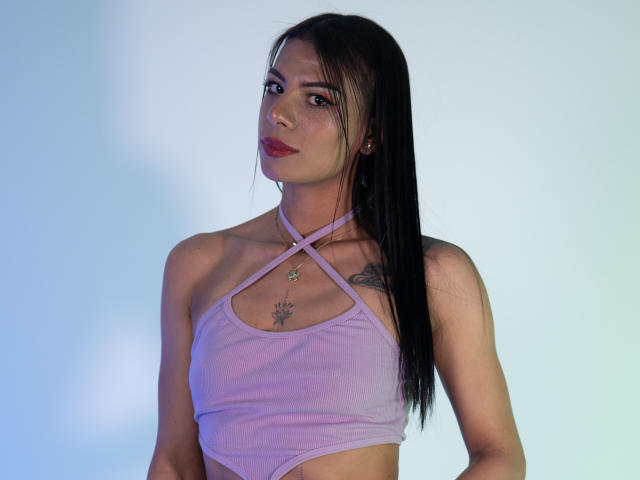 MelissaSpencer with long black hair, wearing a purple halter top, showing small tits, a tattoo on her left shoulder, and a necklace with a star. Light blue background.